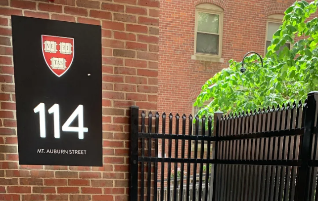 A black sign with white numbering and the Harvard shield that says 114 Mt. Auburn Street, on a brick wall